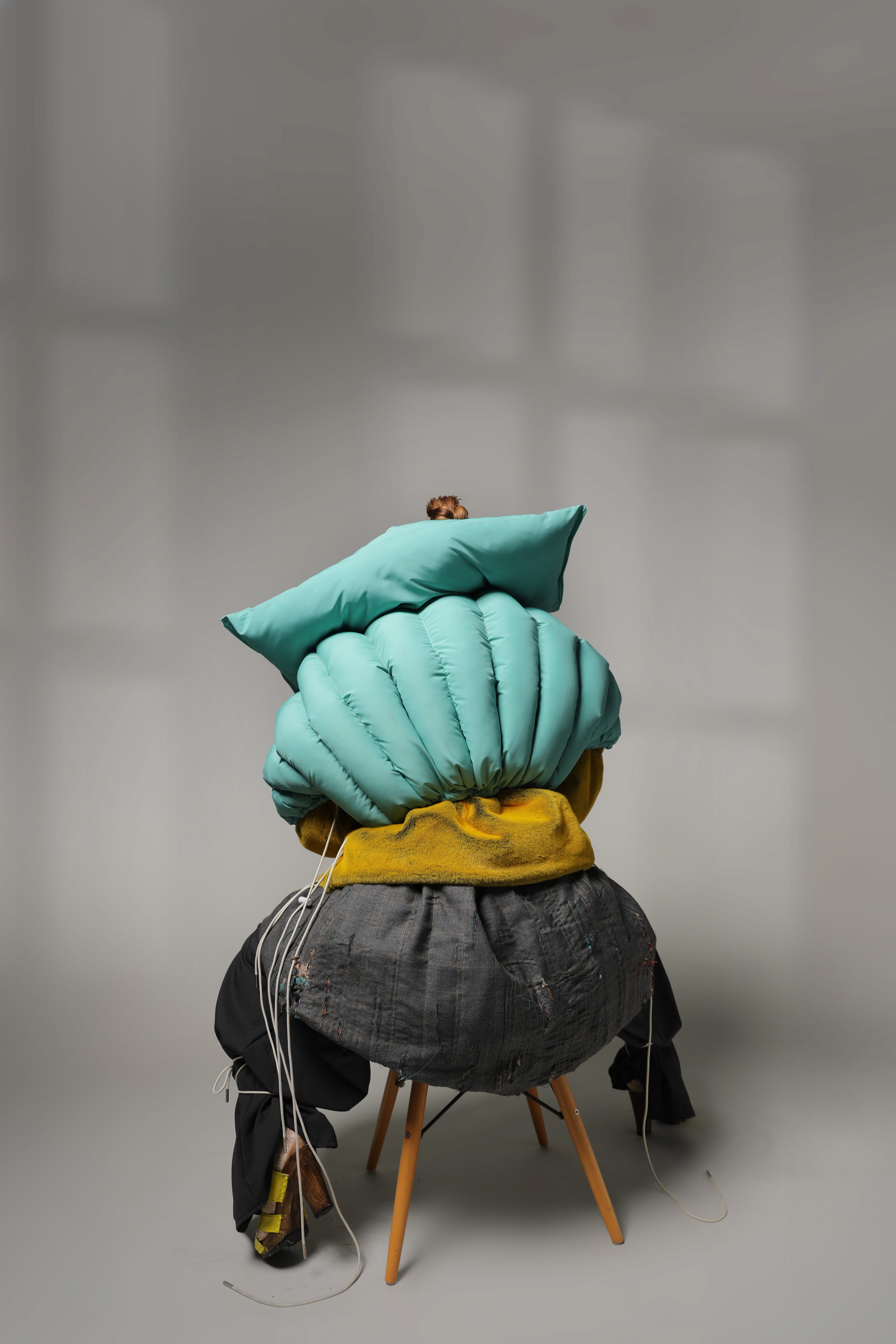 Back view of the seated model almost hidden by the rounded mint puffer, pillow-like collar and ballooning grey hem, on a grey backdrop