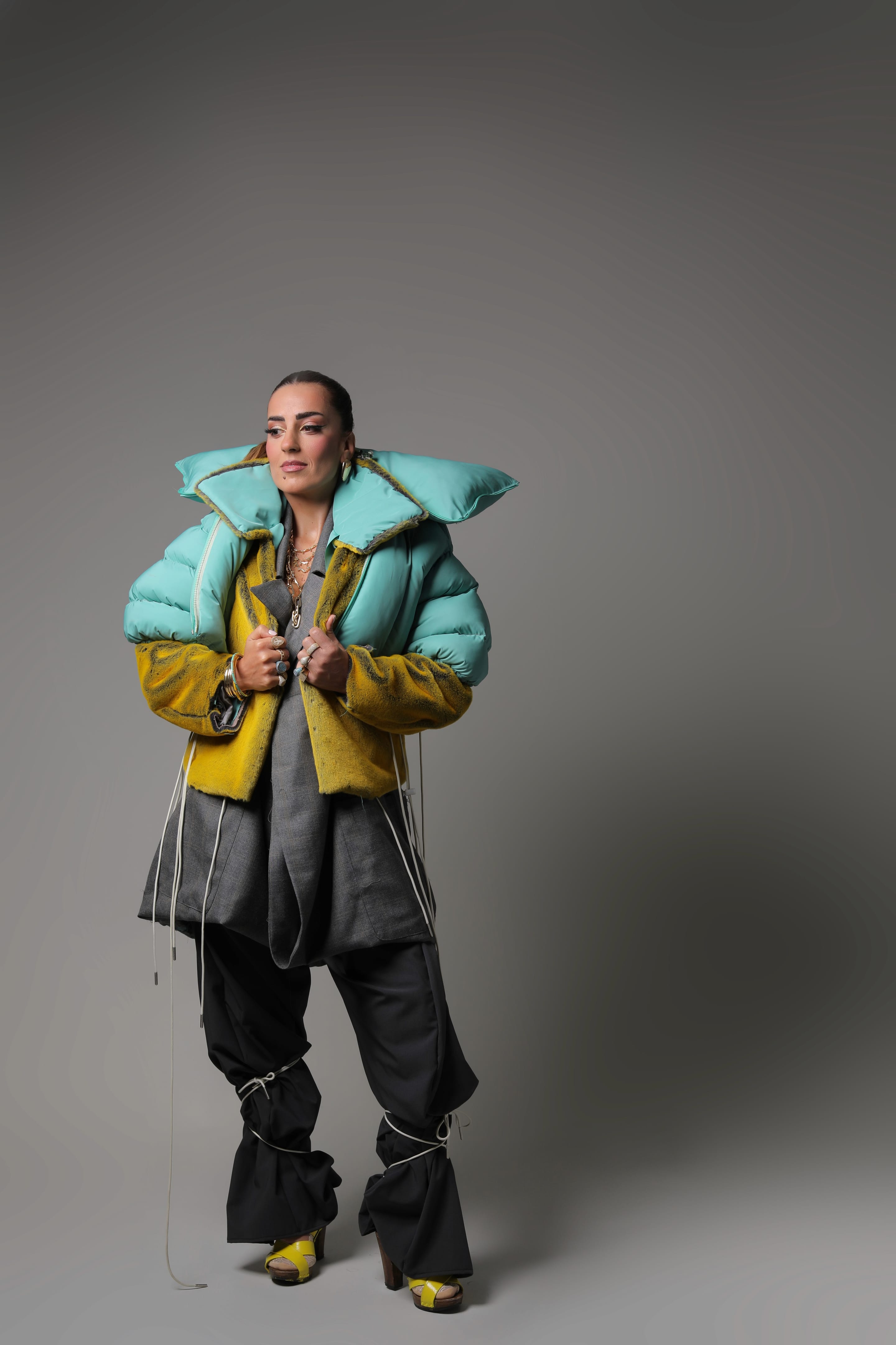 Full-length shot of the model standing and gripping the yellow plush jacket fronts beneath an oversized mint padded collar, looking aside