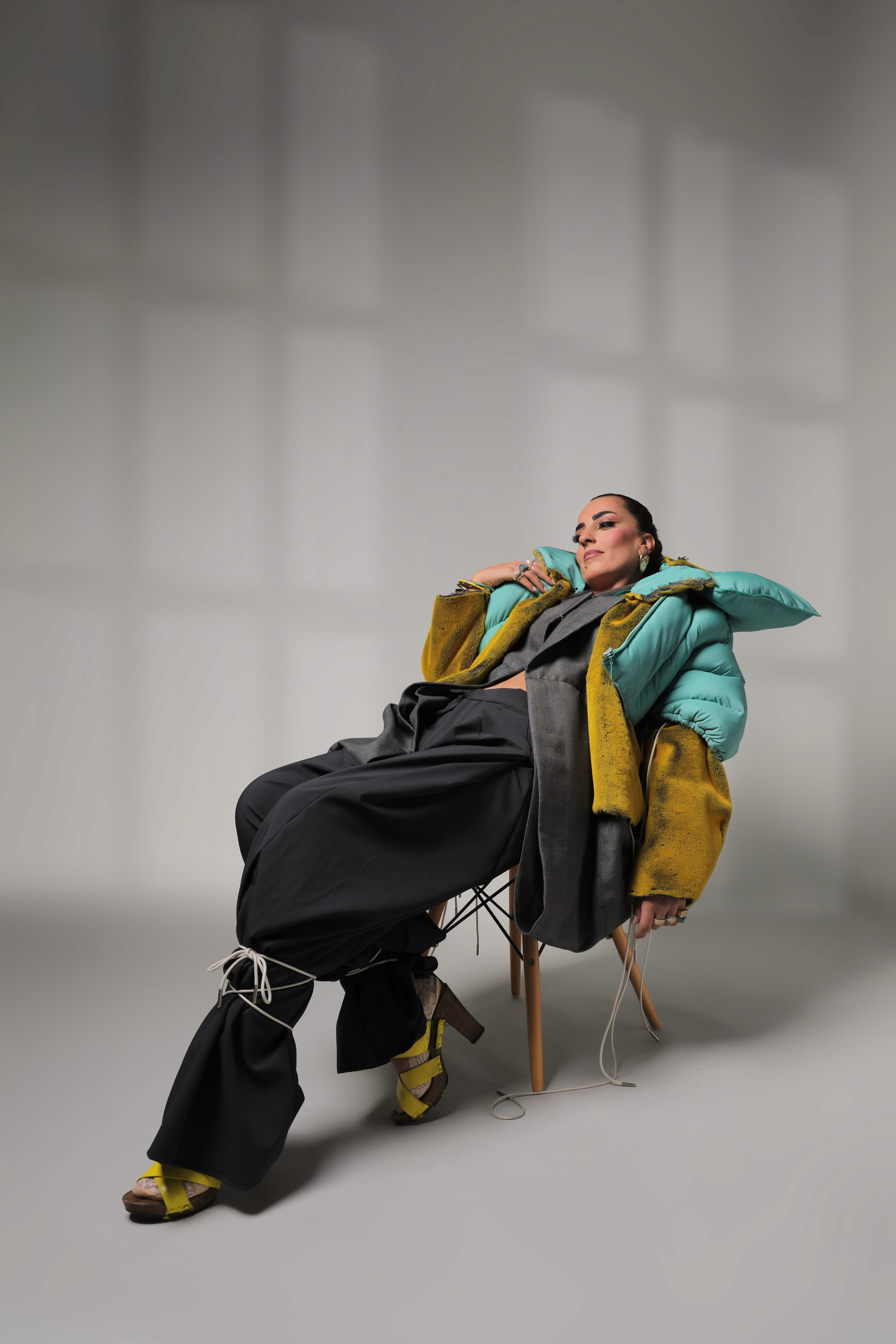 Model reclines on a wooden chair with one leg stretched out, wearing the mint puffer, yellow plush jacket and charcoal cord-tied trousers