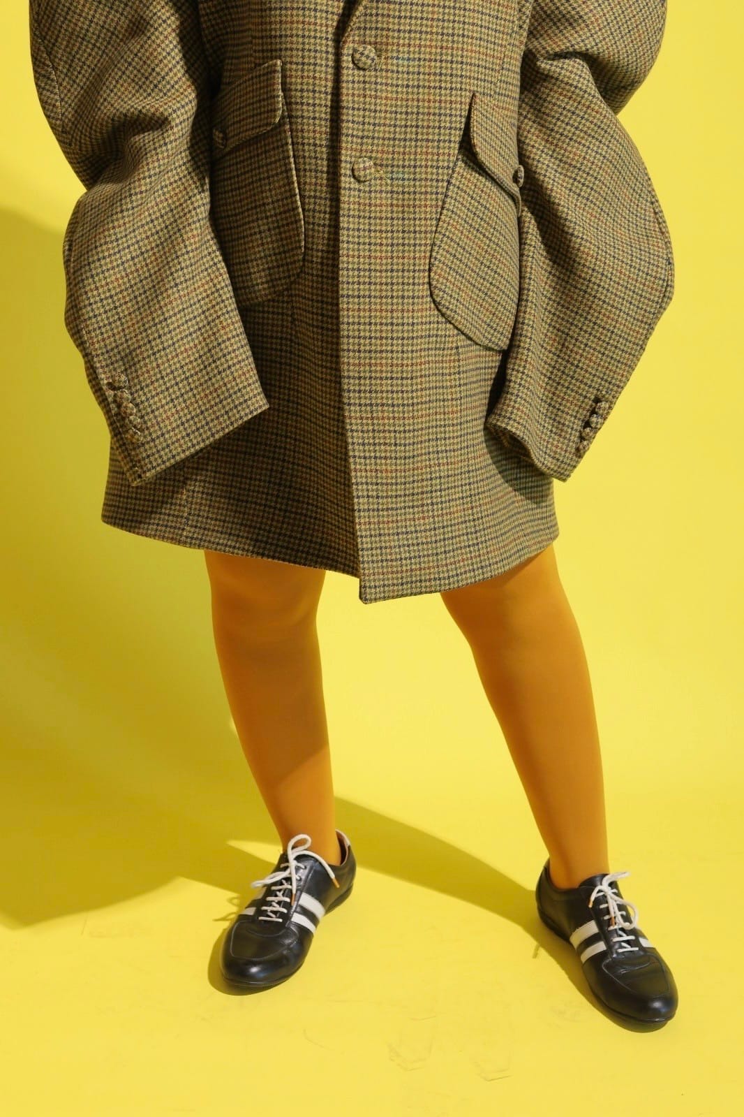 Lower-half crop of an oversized olive houndstooth blazer with flap pockets over mustard tights and black trainers on a yellow backdrop