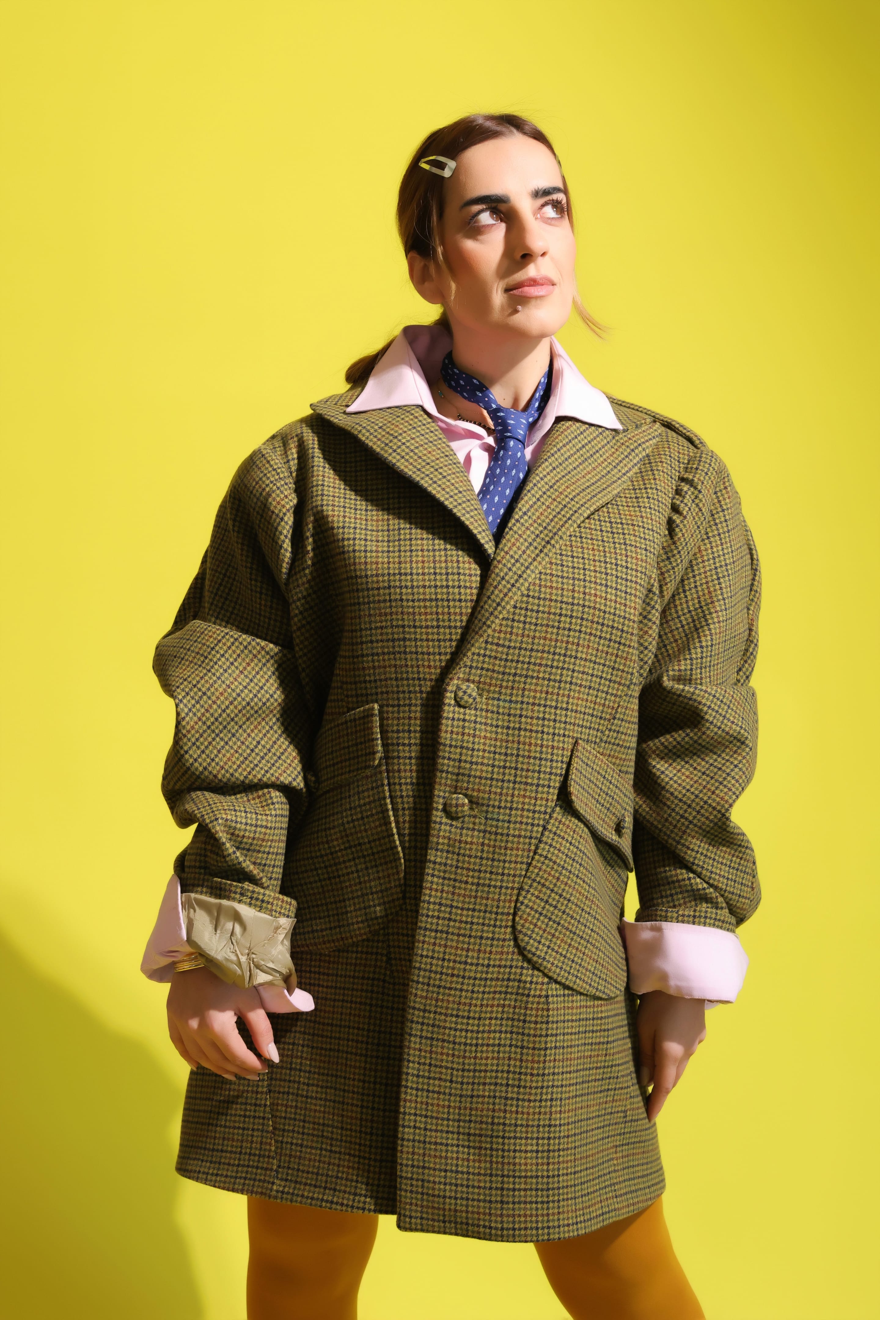 Model in an oversized olive houndstooth blazer over a pink shirt and blue polka-dot tie glances upward against a yellow backdrop