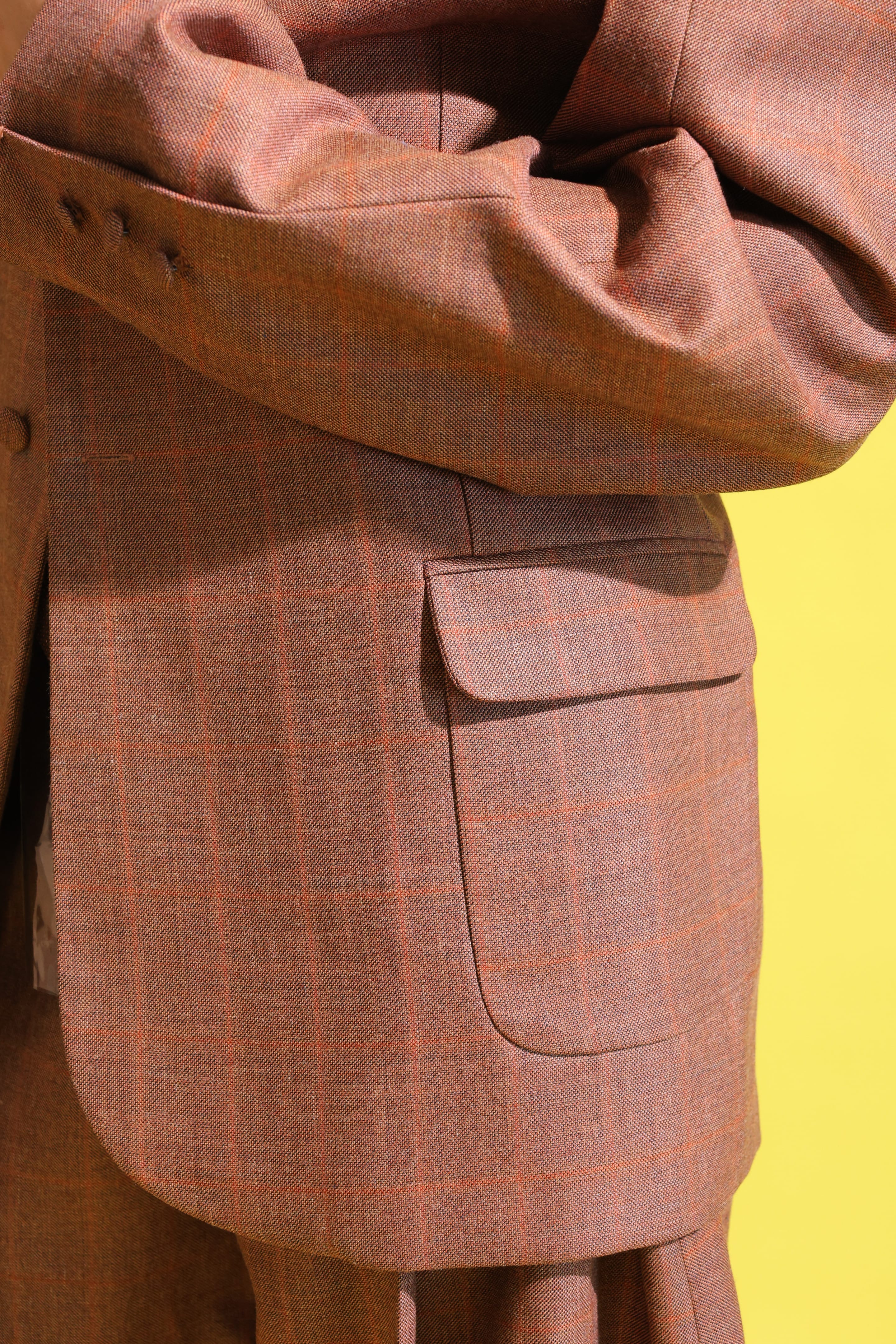 Detail of a rust windowpane-check blazer showing folded arms, cuff buttons and a rounded flap pocket against yellow