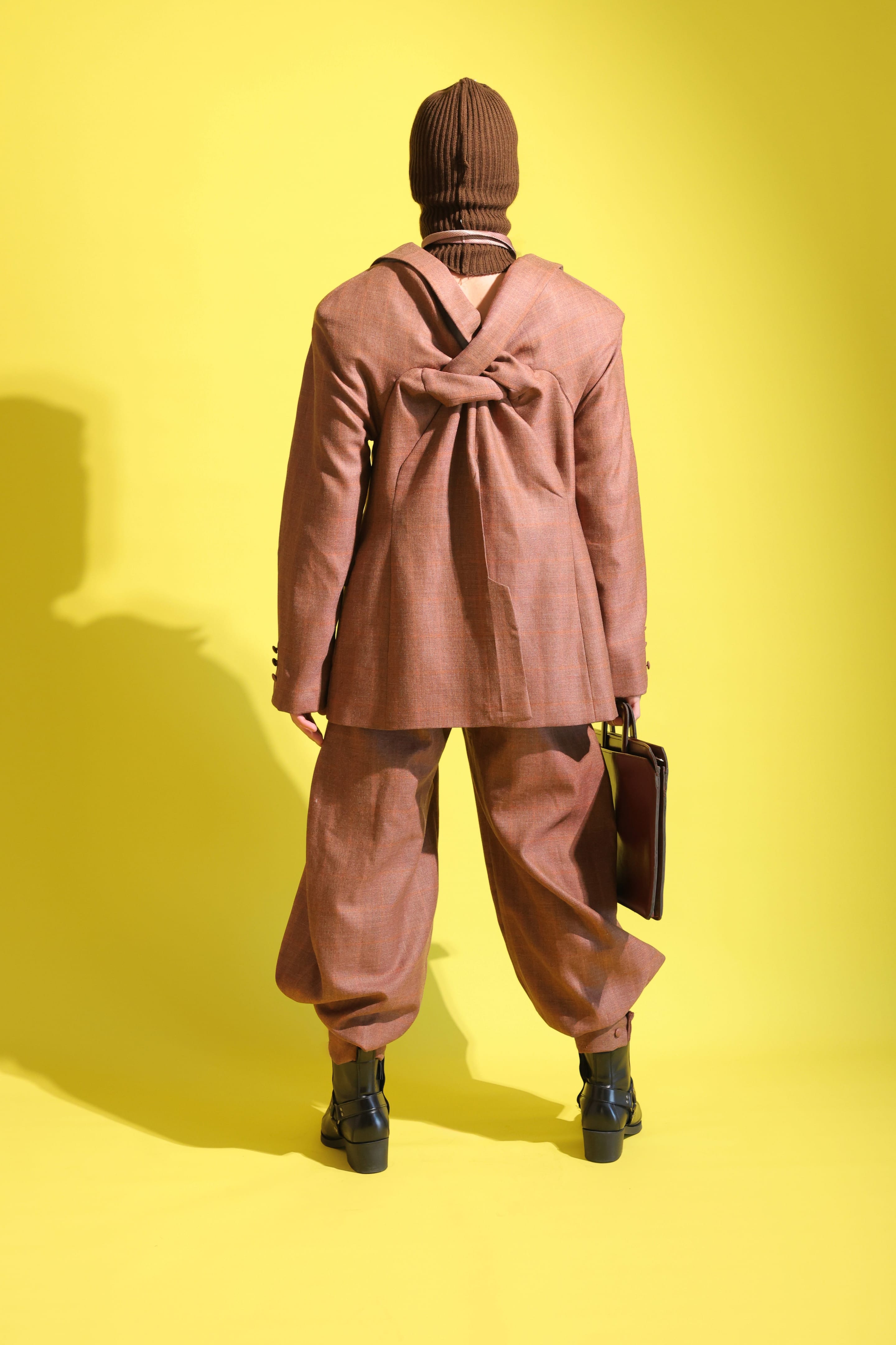 Full-length back view of a rust check suit with a knotted loop across the blazer's back, gathered trousers and black boots