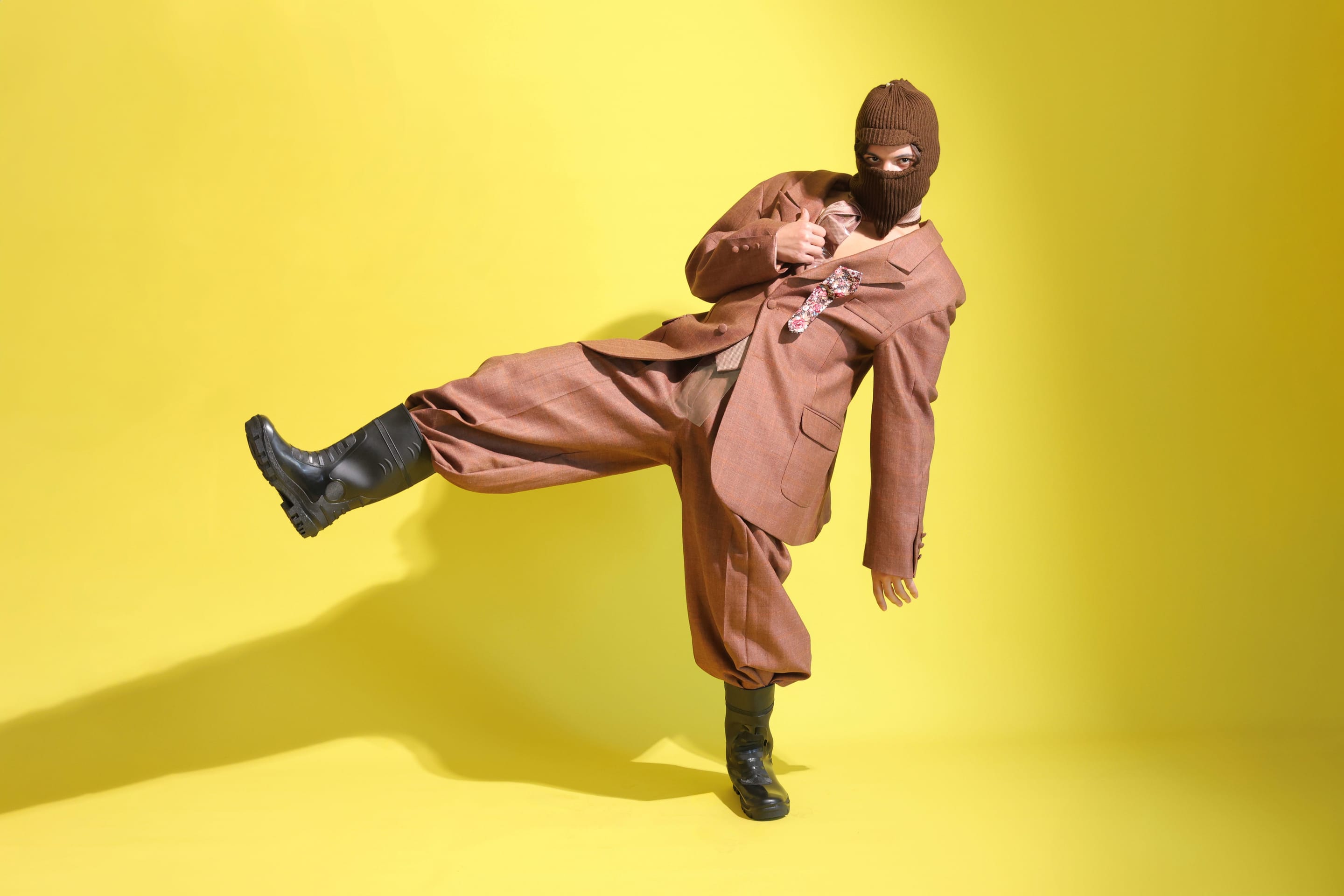 Model in a brown knit balaclava and rust check suit kicks one booted leg high against a bright yellow backdrop