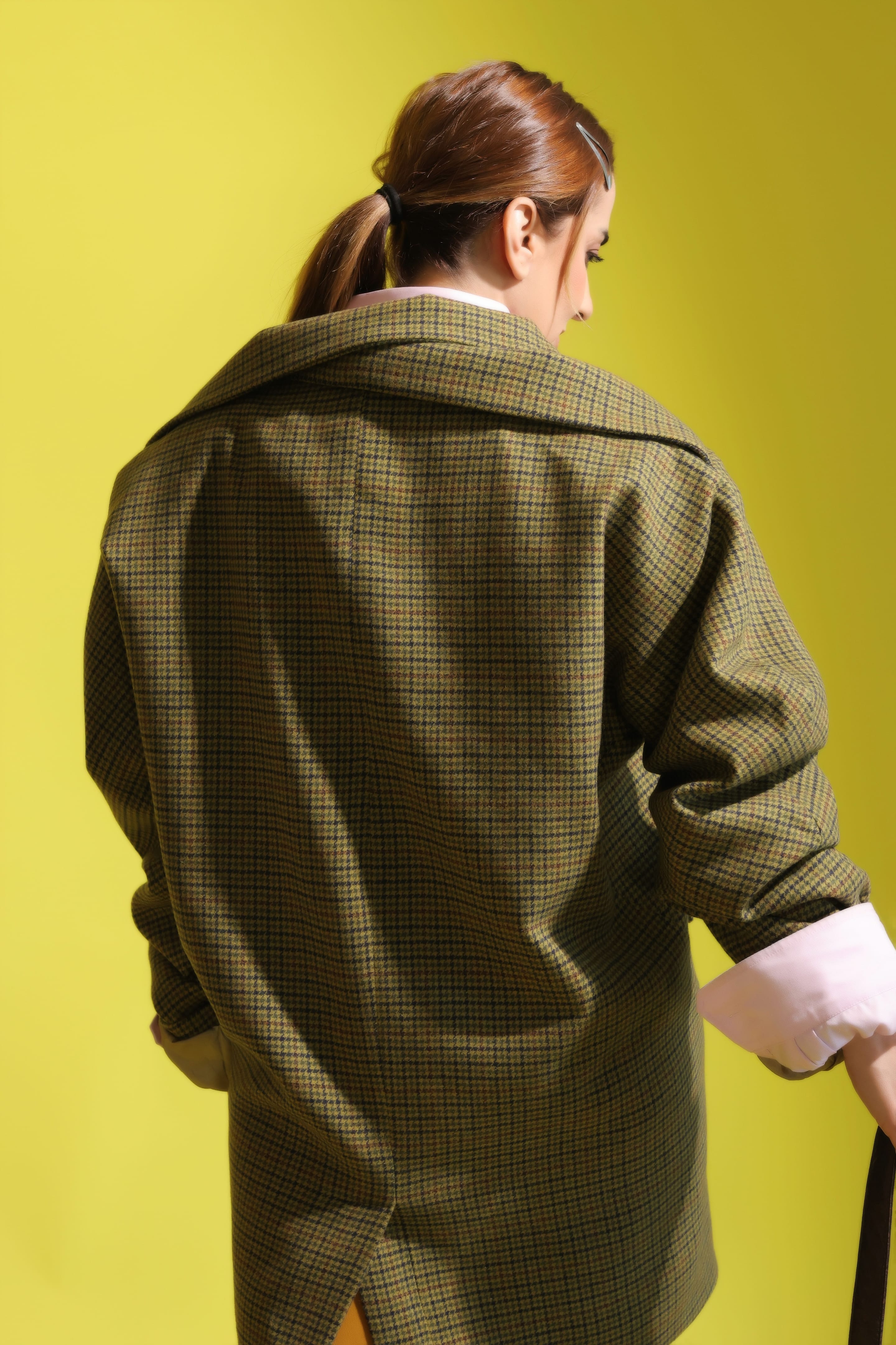 Back view of the model with a ponytail in an oversized olive houndstooth blazer, its broad collar and a pink shirt cuff visible