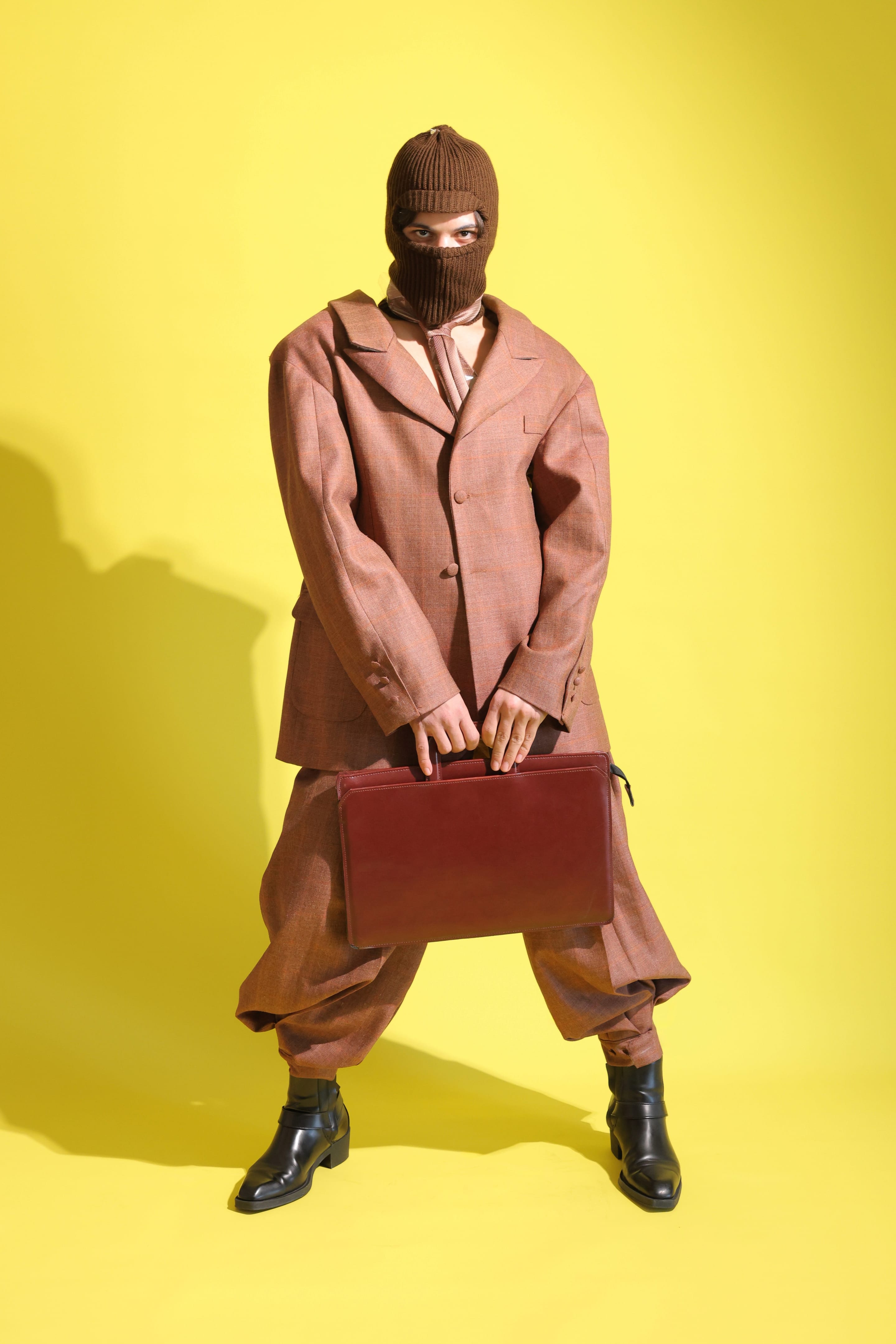 Full-length front view of the model in a brown balaclava and rust check suit, holding a burgundy briefcase with both hands