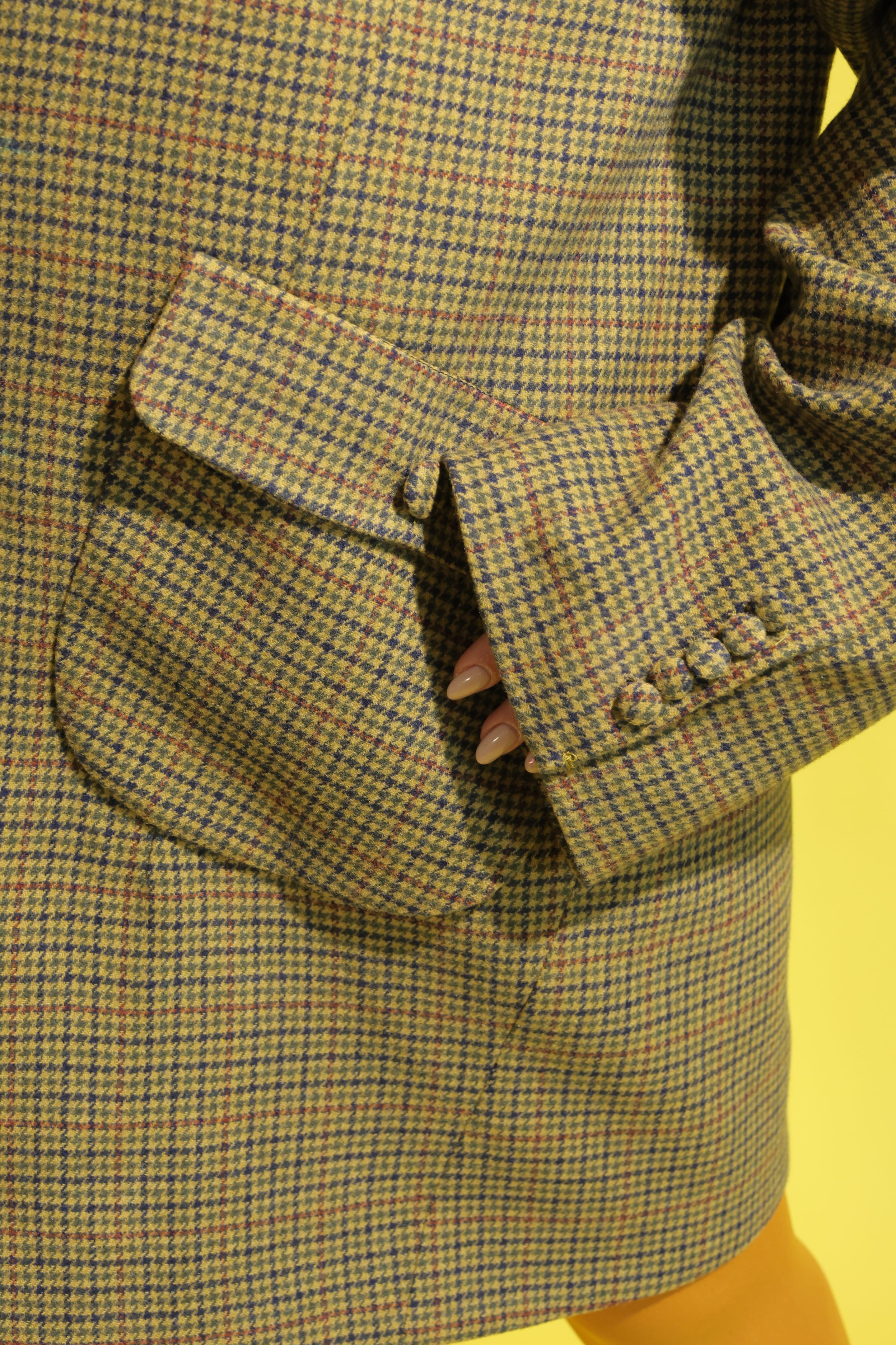 Detail of an olive houndstooth blazer's curved flap pocket and buttoned cuff, fingertips slipped into the pocket, yellow behind