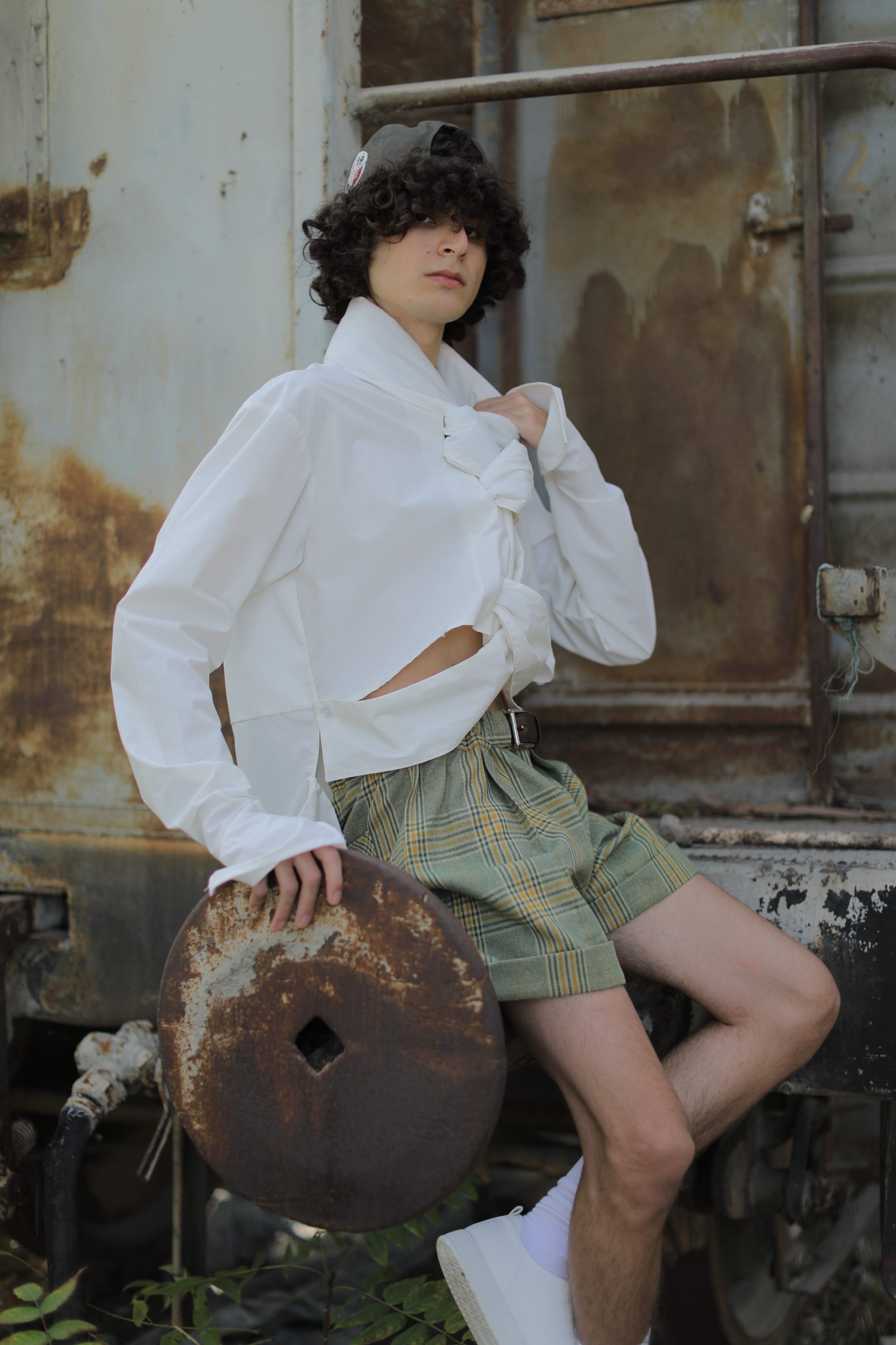 Model in a cropped white shirt with knotted collar and green plaid shorts leans against a rusted train carriage, one knee raised