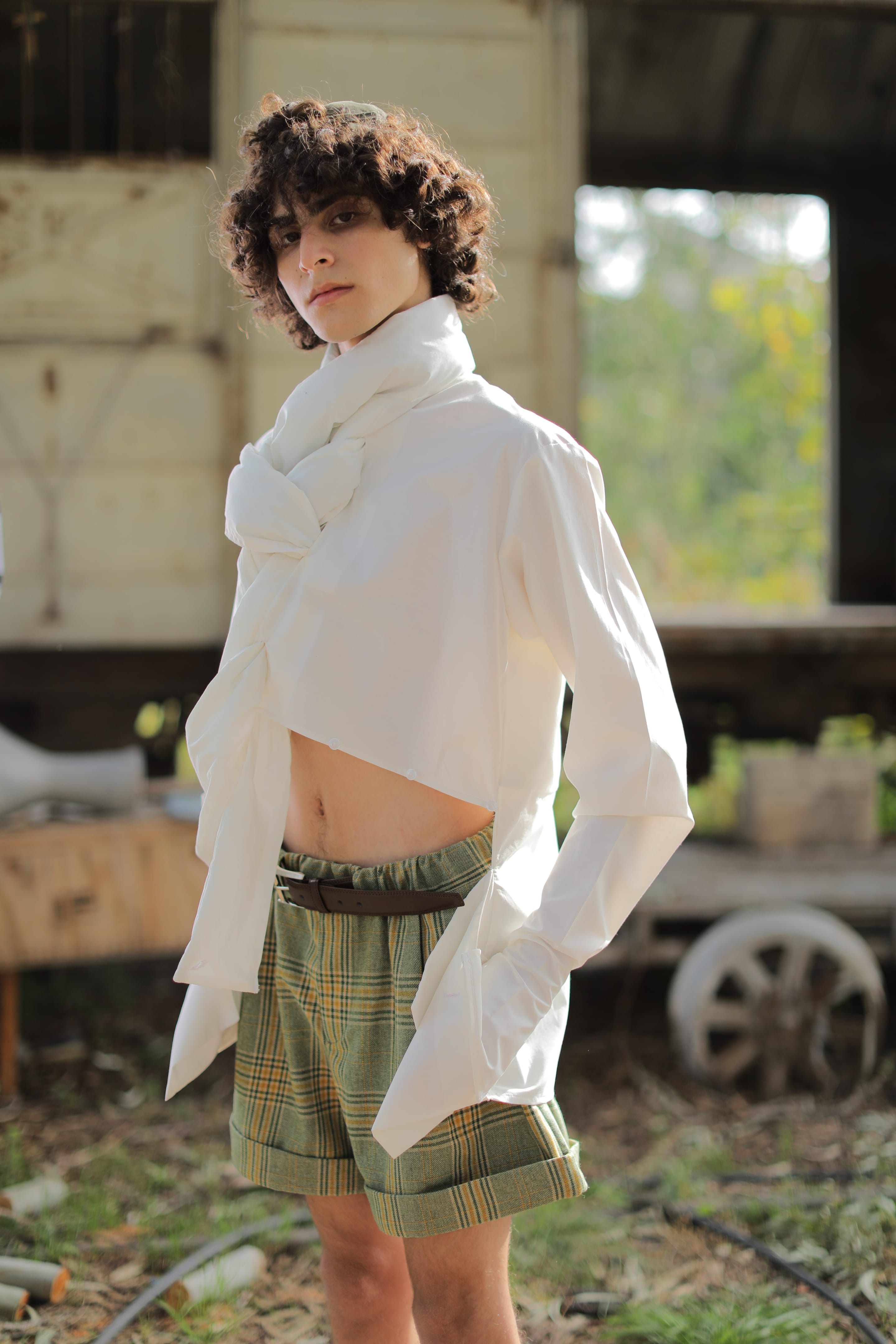 Three-quarter view of the model in a cropped white shirt knotted at the collar and plaid shorts, hand in pocket, cart wheel behind