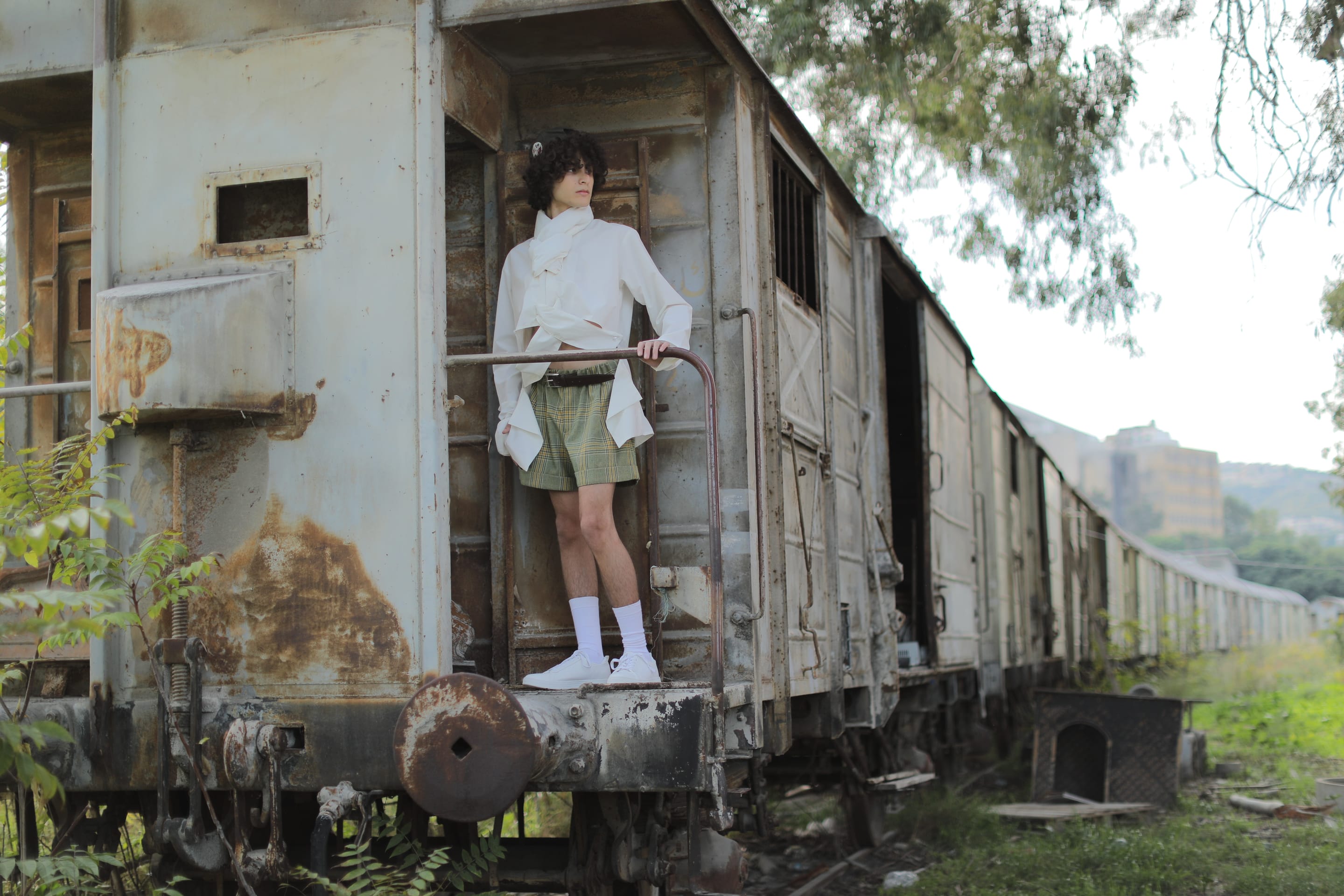 Model stands on the end platform of a rusted train carriage holding the rail, in the white shirt, plaid shorts and white sneakers