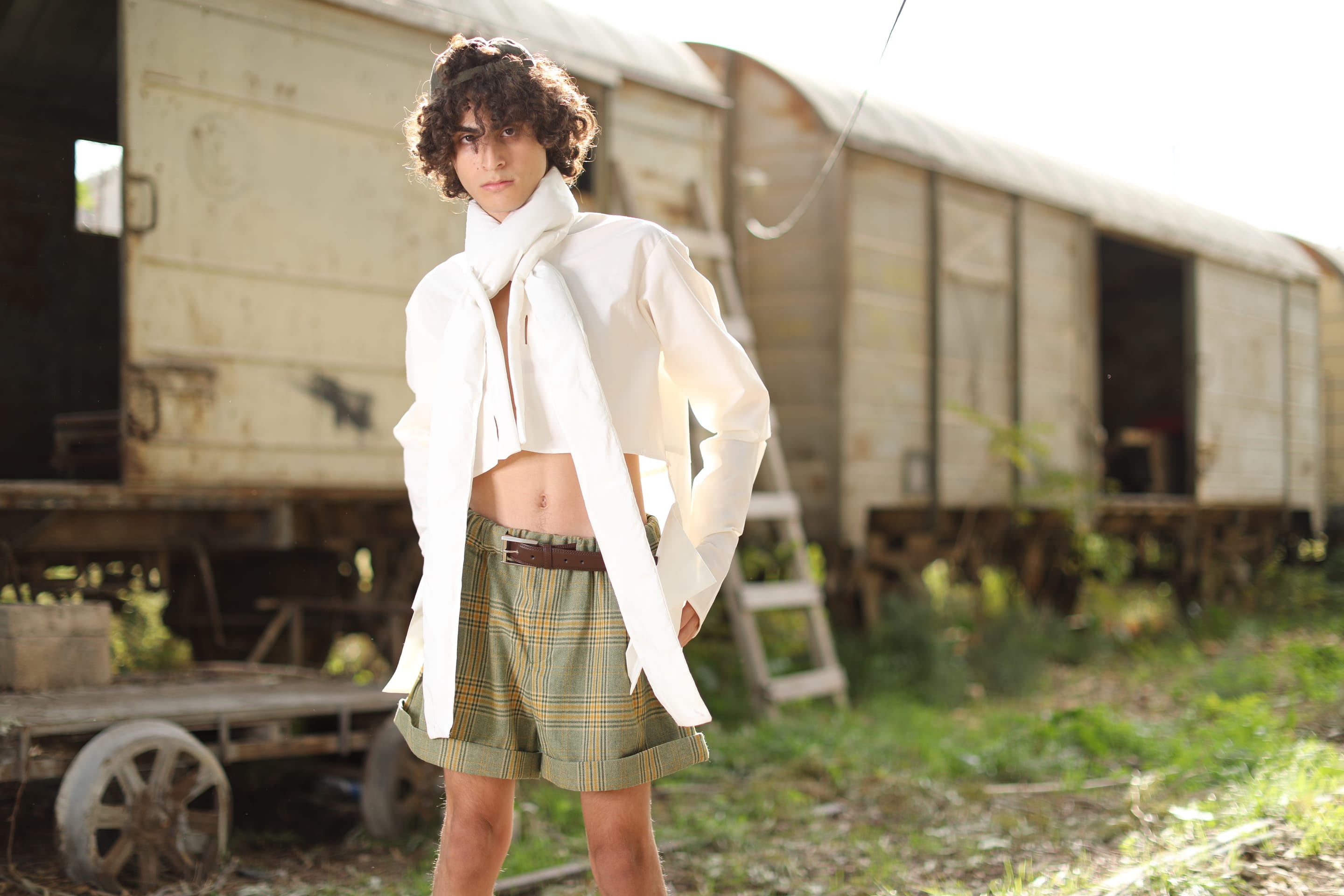 Model stands before weathered freight carriages in a white shirt whose long tie-like collar falls open over a bare midriff and plaid shorts