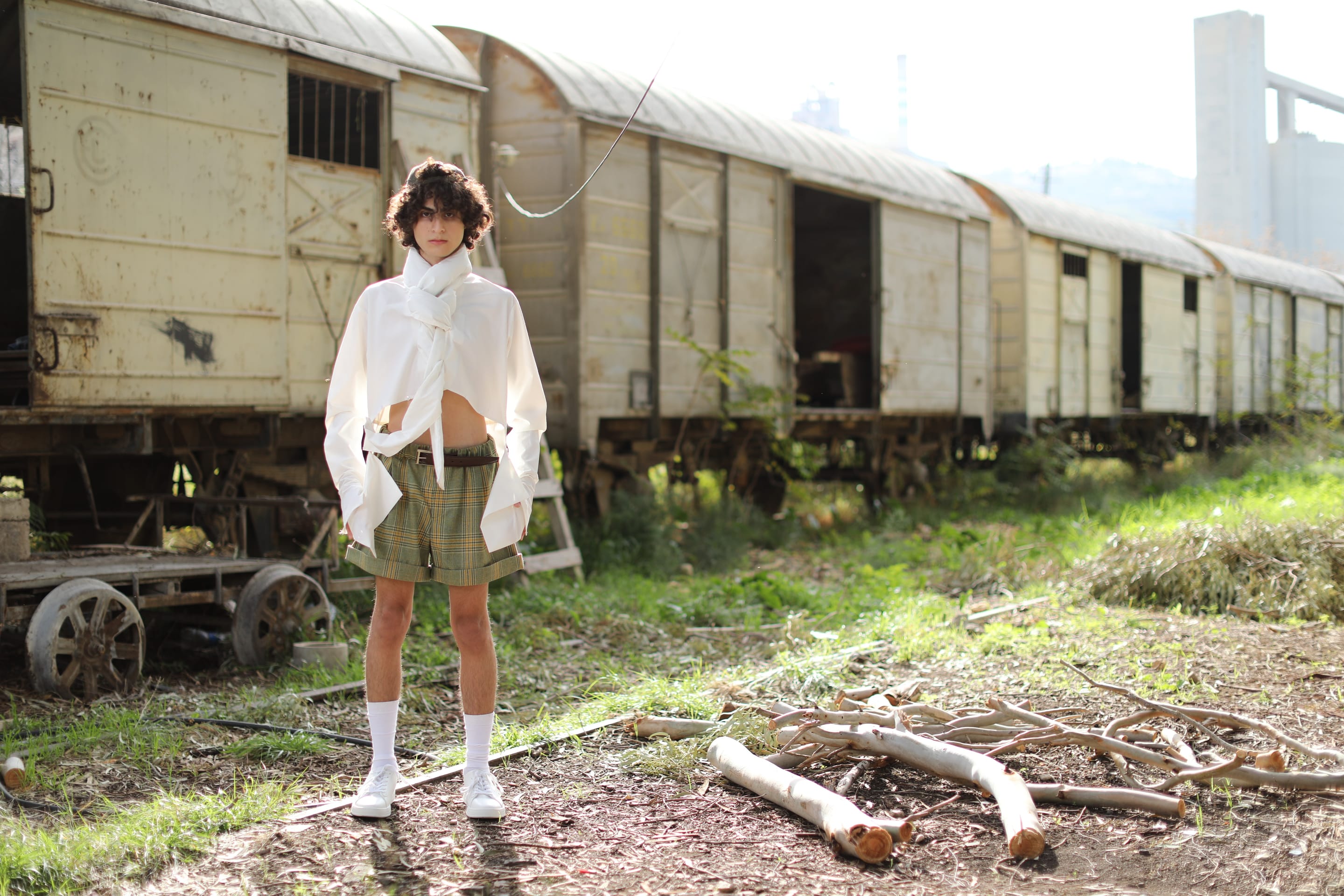 Wide full-length shot of the model in the white shirt and plaid shorts beside a row of weathered freight carriages, cut branches in front