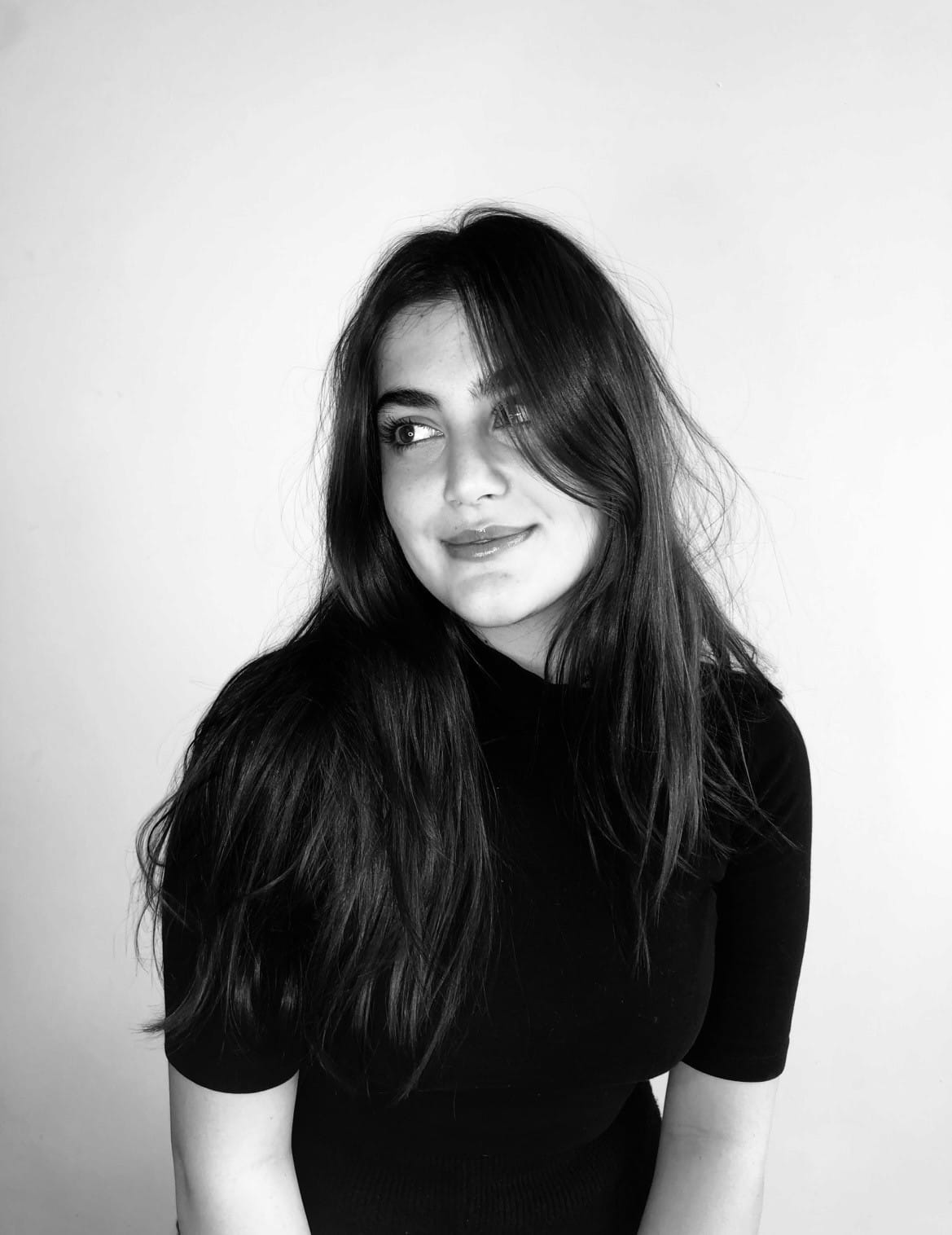 Black-and-white portrait of a smiling person with long dark hair glancing sideways, in a black short-sleeved top against a plain wall